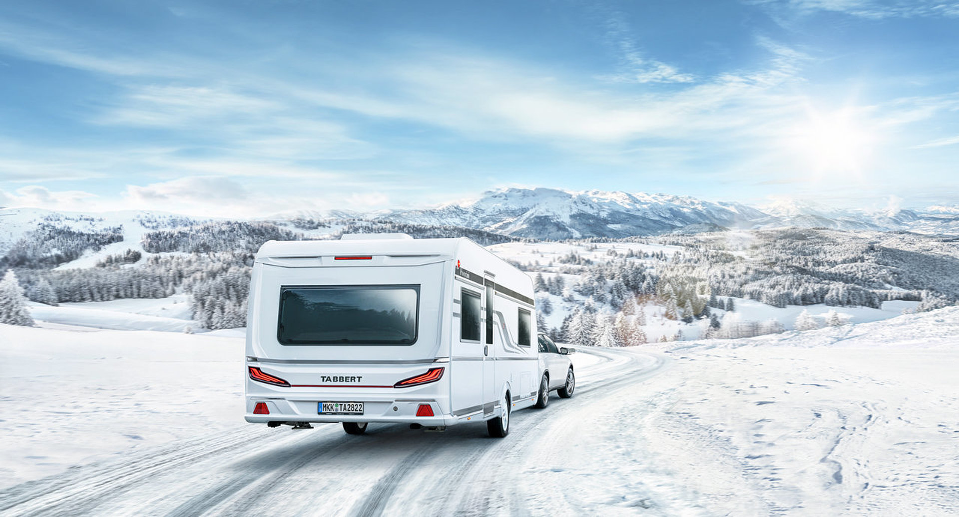 Ein Pkw, der mit einem Wohnwagen durch eine Schneelandschaft fährt.