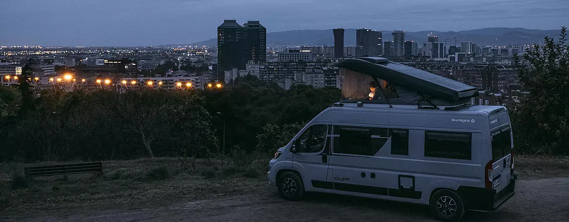 Eine Familie steht im gekauften Wohnmobil im Dunkeln auf einem Berg über einer Skyline.